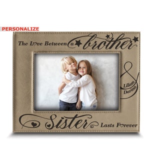 May include: A beige leather photo frame with a stitched border. The frame features a photo of two children hugging, with the text "The Love Between a Brother & Sister Lasts Forever" and the names "Lillian & Darina" inscribed on the frame.