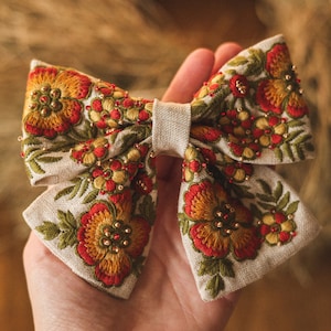 Könnte beinhalten: Eine große, handgefertigte Schleife aus beigefarbenem Leinenstoff mit gestickten roten, orangefarbenen und gelben Blumen und grünen Blättern. Die Schleife ist mit kleinen goldenen und roten Perlen verziert.