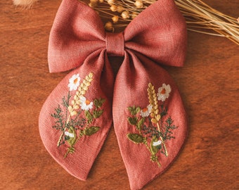 May include: A large, dusty rose-colored bow with embroidered floral details. The bow features white daisies, wheat stalks, and green leaves. The bow is made of linen and is set against a wooden background with dried wheat.