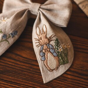 Op de afbeelding: Een beige linnen strik met een geborduurd beeld van Peter Rabbit in een blauw jasje, omringd door groene bladeren en witte bloemen.