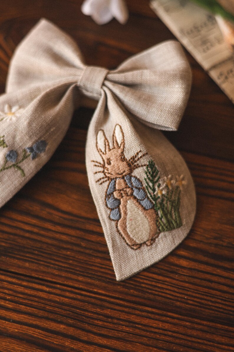 Op de afbeelding: Een beige linnen strik met een geborduurd beeld van Peter Rabbit in een blauw jasje, omringd door groene bladeren en witte bloemen.
