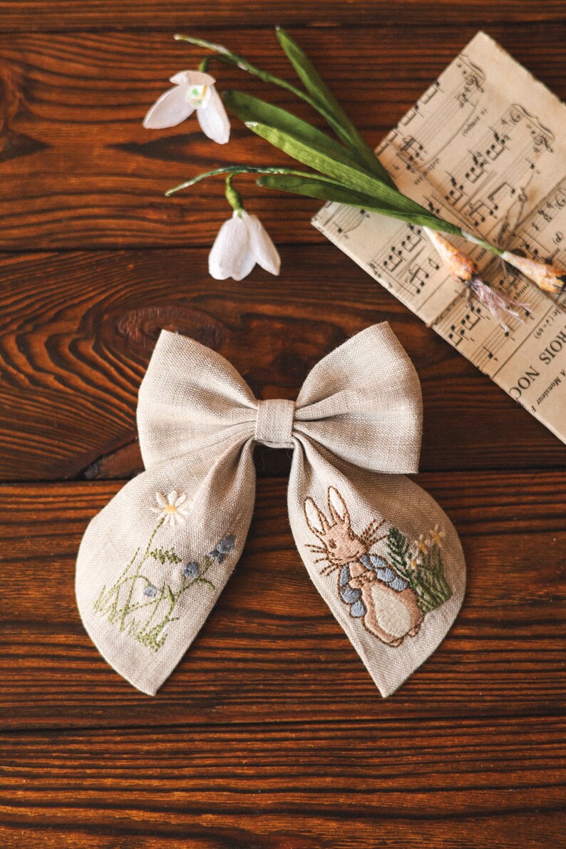 Op de afbeelding: Een beige linnen strik met geborduurde details. De ene kant heeft een bloemenpatroon met madeliefjes en blauwe bellen. De andere kant heeft een Peter Rabbit-personage in een blauw jasje.