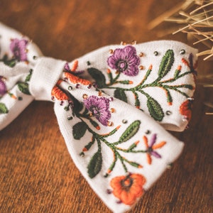 以下が含まれることがあります： 白の生地の蝶ネクタイ。紫、オレンジ、緑色の刺繍の花柄。花と葉の細かい模様で、小さなビーズがアクセント。背景は温かみのある木製の表面。