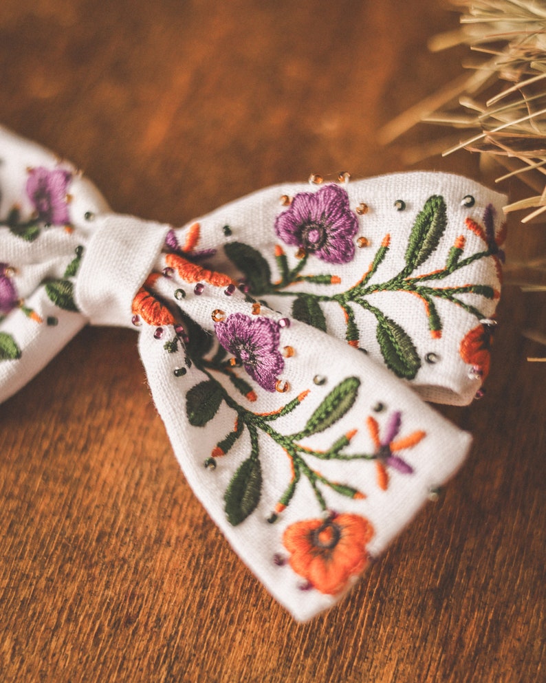 以下が含まれることがあります： 白の生地の蝶ネクタイ。紫、オレンジ、緑色の刺繍の花柄。花と葉の細かい模様で、小さなビーズがアクセント。背景は温かみのある木製の表面。