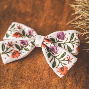 以下が含まれることがあります： オレンジ、紫、緑色の刺繍の花柄が施された白い生地の蝶ネクタイ。 蝶ネクタイは小さな銀色のビーズで装飾され、木製の背景に配置されています。