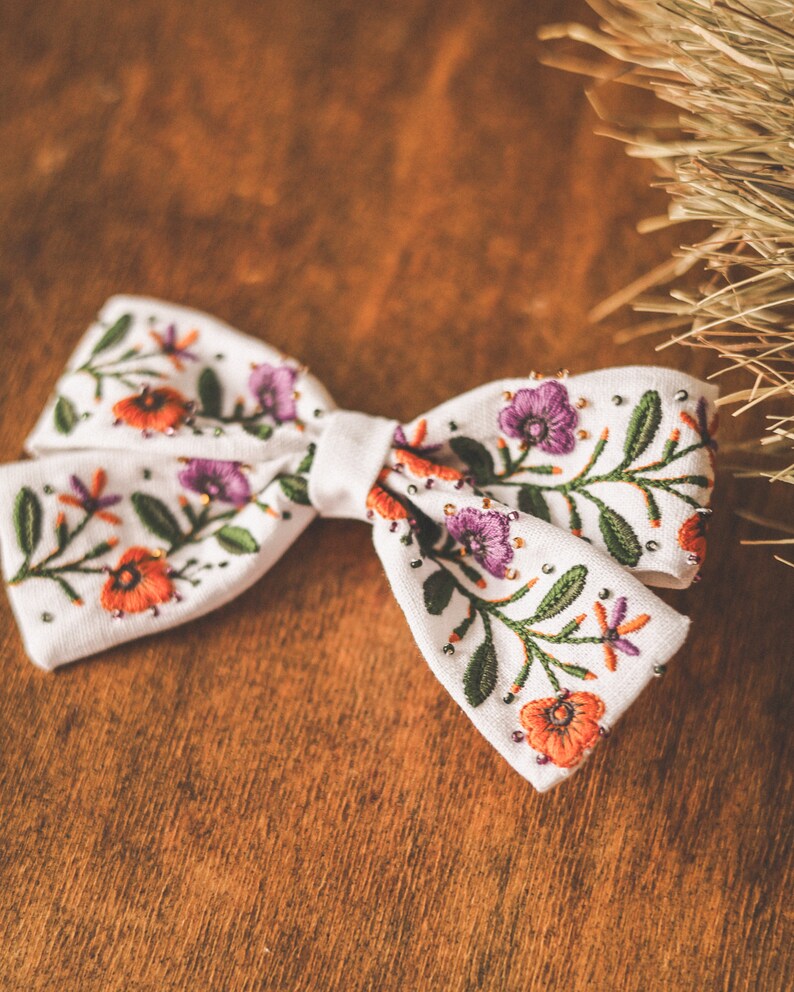 以下が含まれることがあります： オレンジ、紫、緑色の刺繍の花柄が施された白い生地の蝶ネクタイ。 蝶ネクタイは小さな銀色のビーズで装飾され、木製の背景に配置されています。