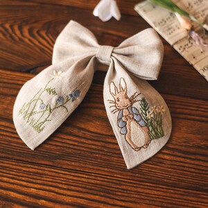 Op de afbeelding: Een beige linnen strik met een geborduurd Peter Rabbit-ontwerp. De strik heeft een bruin konijn dat een blauw jasje draagt en een bloem vasthoudt. De borduurwerk bevat ook groene bladeren en witte bloemen.