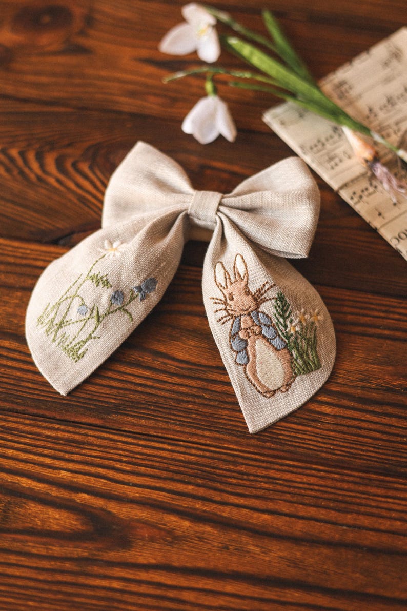 Op de afbeelding: Een beige linnen strik met een geborduurd Peter Rabbit-ontwerp. De strik heeft een bruin konijn dat een blauw jasje draagt en een bloem vasthoudt. De borduurwerk bevat ook groene bladeren en witte bloemen.