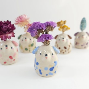 May include: A selection of ceramic dog-shaped bud vases, each holding dried flowers. The vases are off-white with colourful accents, including blue, pink, and brown spots. The flowers are various colours, including purple, pink, and yellow.