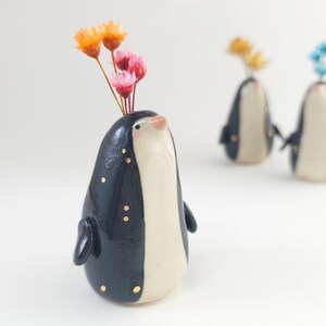May include: A navy blue ceramic penguin vase with a white front and gold detailing. It contains a small arrangement of orange and pink dried flowers. Two other penguin vases are visible in the background.