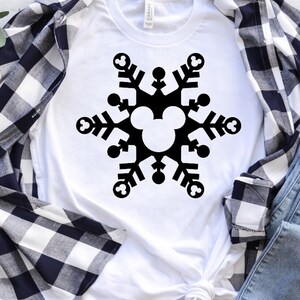 Op de afbeelding: Een wit T-shirt met een zwart sneeuwvlokdesign met een Mickey Mouse-hoofd in het midden. De sneeuwvlok bestaat uit zes punten met Mickey Mouse-oren aan elk punt. Het T-shirt ligt op een wit-blauw geruit flanellen shirt en een blauwe spijkerbroek.