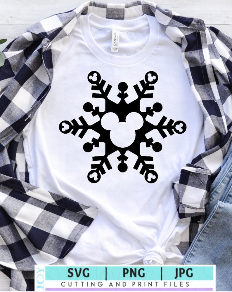 Op de afbeelding: Een wit T-shirt met een zwart sneeuwvlokdesign met een Mickey Mouse-hoofd in het midden. De sneeuwvlok bestaat uit zes punten met Mickey Mouse-oren aan elk punt. Het T-shirt ligt op een wit-blauw geruit flanellen shirt en een blauwe spijkerbroek.