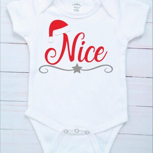 May include: White baby bodysuit with a red Santa hat and the word "Nice" in red script. A gray star and decorative line are below the word. The bodysuit has short sleeves and snaps at the bottom.