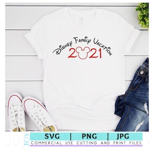 May include: White t-shirt with black text that reads "Disney Family Vacation 2021" with a red Mickey Mouse head in between the numbers. The shirt is laying on a white wooden surface with a pair of white sneakers and blue jeans.