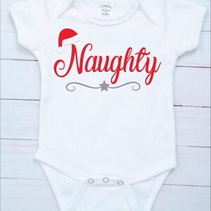 May include: White baby onesie with a red Santa hat and the word "Naughty" in red script. A gray decorative line with a star is below the word. The onesie has short sleeves and snap closures.