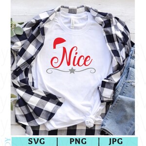 May include: White t-shirt with the word "Nice" in red script, topped with a Santa hat. A gray star and decorative line are below the word. The shirt is layered with a black and white plaid shirt and blue jeans.