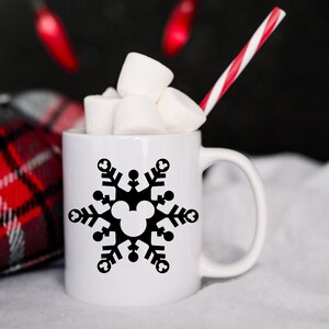 Op de afbeelding: Een witte keramische mok met een zwart sneeuwvlokkenontwerp met een Mickey Mouse-silhouet. De mok is gevuld met marshmallows en heeft een rood-wit gestreepte rietje.
