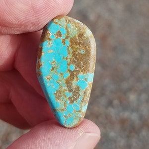 May include: A turquoise cabochon with brown matrix. The turquoise is a bright blue colour and the matrix is a dark brown colour. The cabochon is a teardrop shape.
