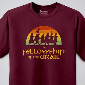 May include: Maroon t-shirt with a graphic of silhouetted figures walking under a sunset. The text "the Fellowship of the Grail" is printed in gold below the graphic. The shirt has a crew neck and short sleeves.