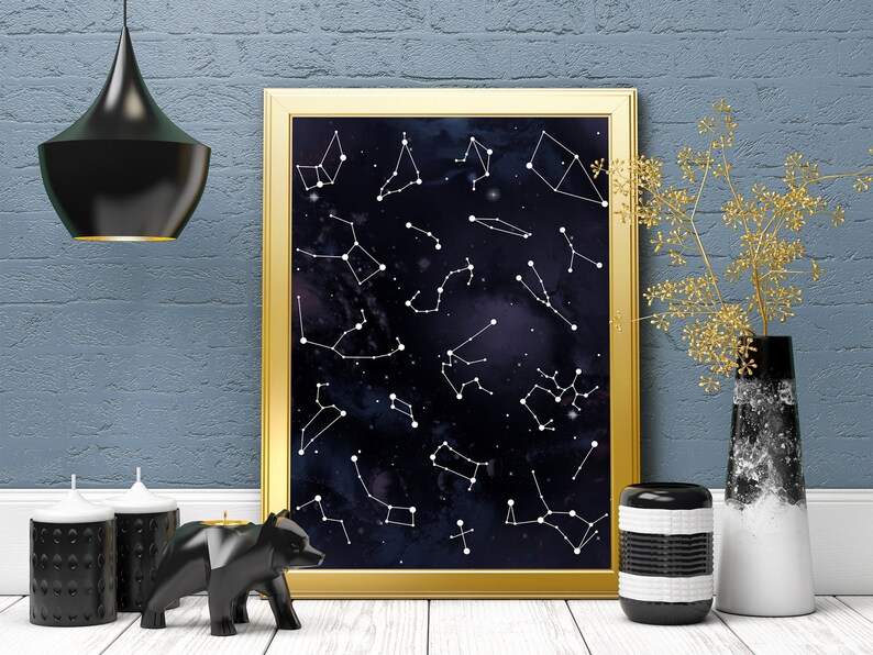 May include: A gold framed print with a black background and white constellations. The print is hanging on a blue brick wall with a black pendant light above it. There are two black candles and a black bear figurine on a white surface in front of the print.