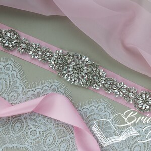 May include: A pink satin ribbon with a crystal embellished belt buckle. The buckle is made of silver-toned metal and features a cluster of clear rhinestones.