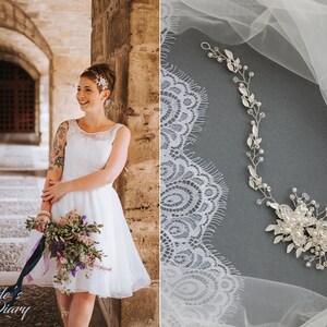 May include: A bride wearing a white lace dress with a floral bouquet and a silver hairpiece. The bride is standing in a stone archway. The image also shows a silver hairpiece with rhinestones and leaves on a white lace fabric.