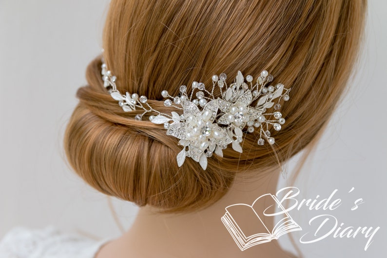May include: A silver and pearl bridal hair comb with a floral design. The comb is worn in a woman's hair, which is styled in a bun.