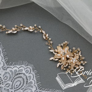 May include: Gold bridal hair vine with delicate leaves and pearl accents. The vine features a larger floral design with pearls and rhinestones. The vine is displayed on a gray background with a piece of white lace and tulle.