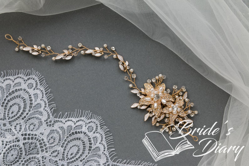 May include: Gold bridal hair vine with delicate leaves and pearl accents. The vine features a larger floral design with pearls and rhinestones. The vine is displayed on a gray background with a piece of white lace and tulle.