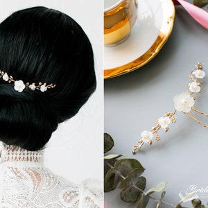 May include: Gold hair comb with white flower accents. The comb is shown in a model's hair and also laid flat on a gray surface.