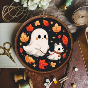 May include: A cross-stitch embroidery hoop with a black background featuring a white ghost holding a leash attached to a white dog. The ghost and dog are surrounded by orange and yellow leaves.