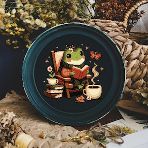 Può includere: Una rana verde con un maglione rosso è seduta su una sedia di legno a leggere un libro. La rana è circondata da fiori, una tazza di tè e una pila di libri. La scena si svolge su uno sfondo blu scuro.