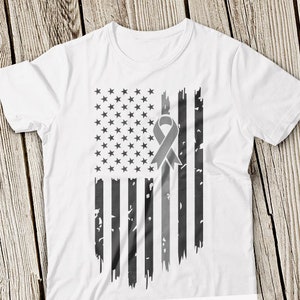 May include: White t-shirt featuring a distressed American flag design in shades of gray. The flag includes stars and stripes, with a gray awareness ribbon overlaying the stripes. The shirt is made of a soft material.
