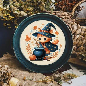 May include: A cross-stitch pattern featuring a cute orange cat wearing a witch's hat and holding a wand. The cat is standing next to a cauldron and a stack of books. The background is a cream color with fall leaves and stars.