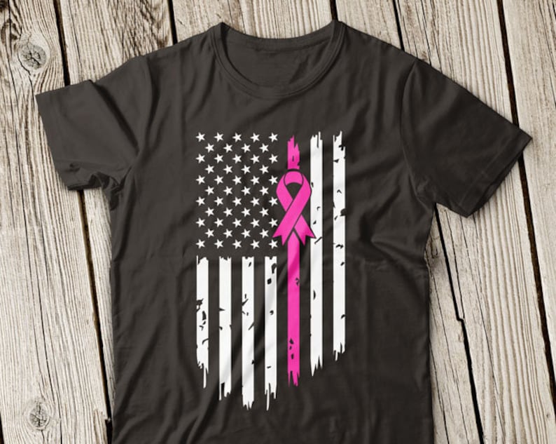 Op de afbeelding: Een zwart T-shirt met een verweerd Amerikaans vlagontwerp met een roze lint in het midden. Het lint is een symbool voor bewustwording van borstkanker.