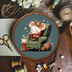 Puede incluir: Un patrón de punto de cruz de un lindo ratón con una diadema floral, sentado en un sillón con un libro y una taza de té. El ratón está rodeado de una bola de hilo y agujas de tejer.