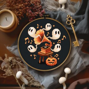 May include: A cross-stitch embroidery hoop featuring a Halloween design. The design includes pixelated ghosts, musical notes, and a jack-o'-lantern around a vintage gramophone. The background is black, and the hoop is set against a neutral fabric.