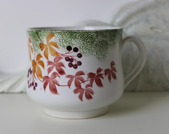Puede incluir: Taza de cerámica blanca con asa, decorada con un diseño de temática otoñal. El diseño presenta hojas naranjas, rojas y amarillas, junto con bayas moradas oscuras, sobre un fondo verde moteado.