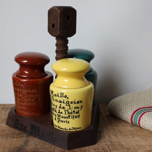 Puede incluir: Un juego de tres tarros de cerámica para condimentos en marrón, amarillo y verde azulado, con un dispensador de tapa de rosca de madera. El tarro amarillo tiene texto negro. El juego se asienta sobre una base de madera oscura. Un paño beige doblado con rayas rojas y verdes está a la derecha.