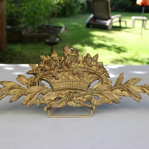 Puede incluir: Una pieza decorativa de metal dorado con un diseño floral. La pieza presenta una cesta de flores con hojas y enredaderas que se extienden a ambos lados.