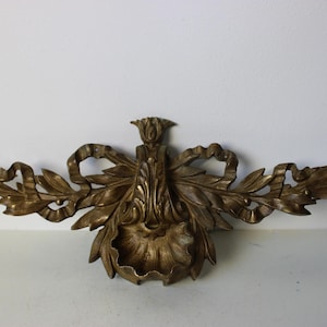Puede incluir: Un soporte de pared decorativo de latón con un diseño floral y un lazo. El soporte tiene un diseño detallado y ornamentado con hojas y volutas.