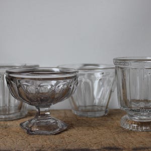Puede incluir: Cuatro vasos de vidrio transparente de diferentes formas y tamaños están dispuestos sobre una superficie de madera. La colección incluye dos vasos cortos y anchos, una copa de postre con pie y un vaso pequeño con pie.