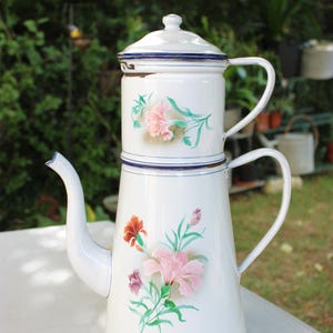 May include: A white enamel coffee pot with a stacked design, featuring a removable top section and a lid. The exterior is decorated with painted floral motifs in shades of pink, orange, and purple with green leaves, accented by thin blue trim.