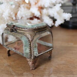 May include: A small, clear glass box with a gold metal frame and ornate details. The box has a hinged lid and sits on four decorative feet.