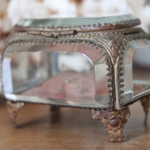 May include: A small, clear glass trinket box with a gold-toned metal frame and ornate detailing. The box has a hinged lid and sits on four decorative feet.