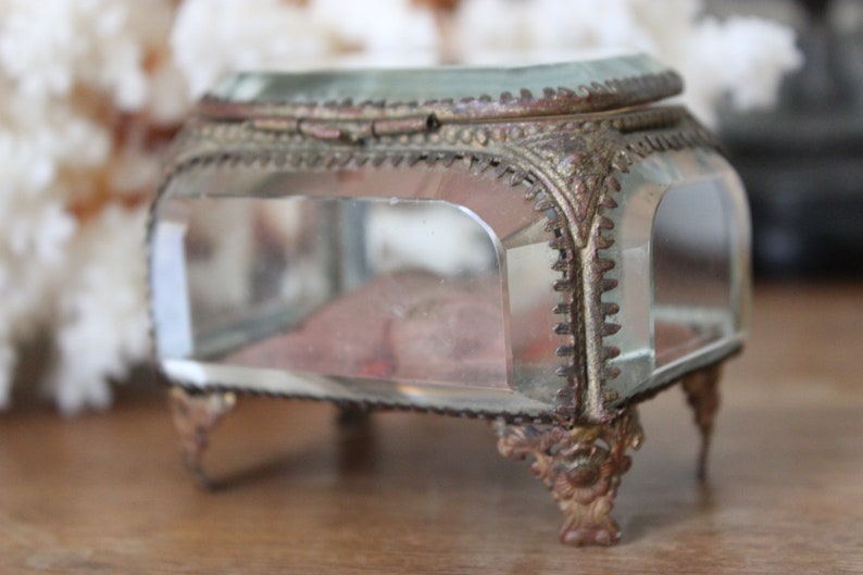 May include: A small, clear glass trinket box with a gold-toned metal frame and ornate detailing. The box has a hinged lid and sits on four decorative feet.