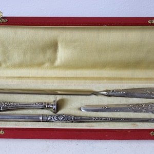 Op de afbeelding: Een set zilveren schrijfgerei in een rode fluwelen doos. De set bevat een briefopener, een pen, een zegel en een klein mes.