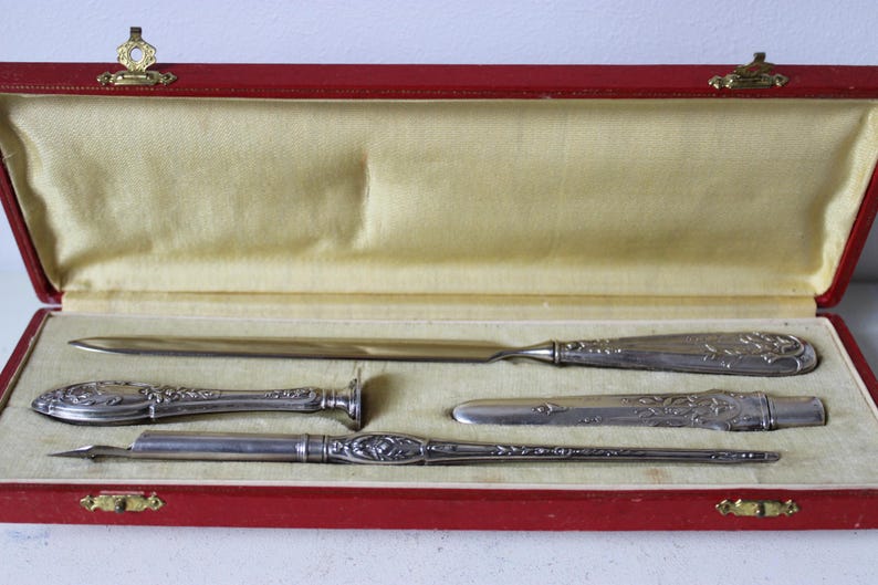 Op de afbeelding: Een set zilveren schrijfgerei in een rode fluwelen doos. De set bevat een briefopener, een pen, een zegel en een klein mes.