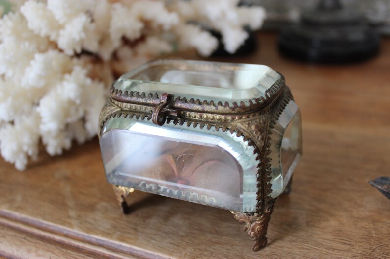 May include: A small, clear glass box with a gold metal trim and feet. The box has a hinged lid with a metal clasp.