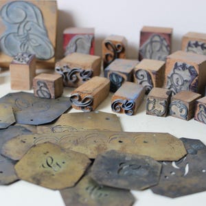 May include: A collection of vintage wooden printing blocks with various designs and letters. The blocks are arranged on a white surface with several metal plates with engraved designs.